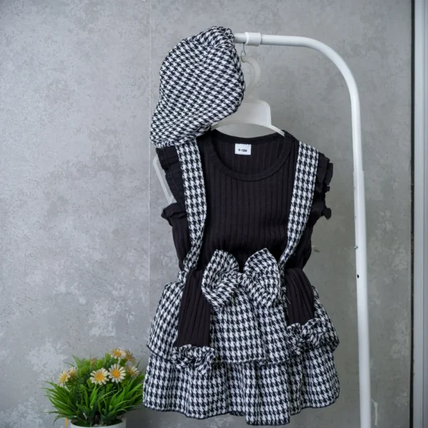 Baby girl’s 3-piece outfit with black ribbed top, houndstooth suspender skirt, large bow, and matching cap on hanger.