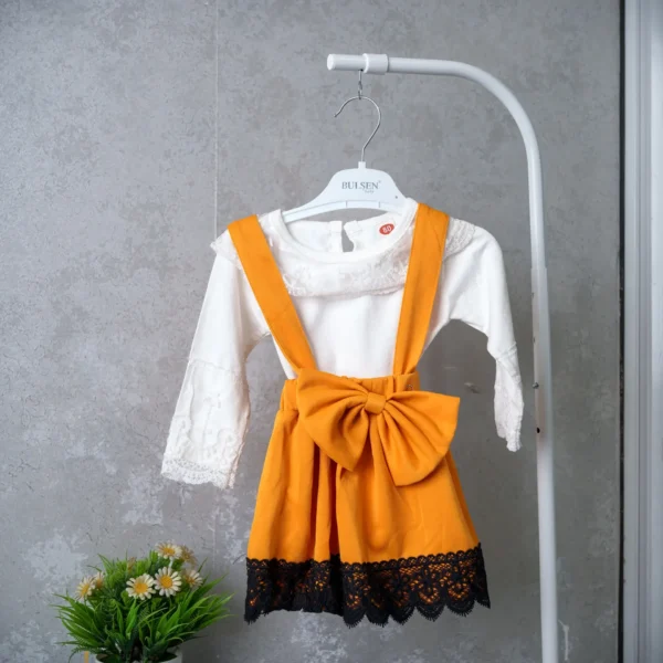 Toddler girl’s white lace long-sleeve top with mustard-yellow suspender skirt, oversized bow, and black lace trim.