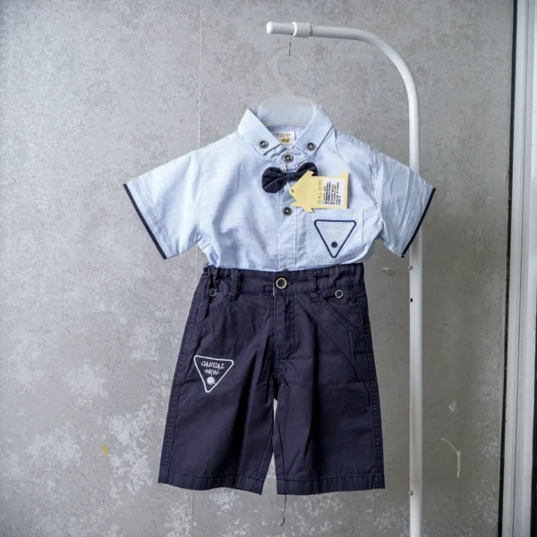 Baby boy’s blue short-sleeve shirt with navy shorts, bow tie, and triangle patch accents, hanging on display.