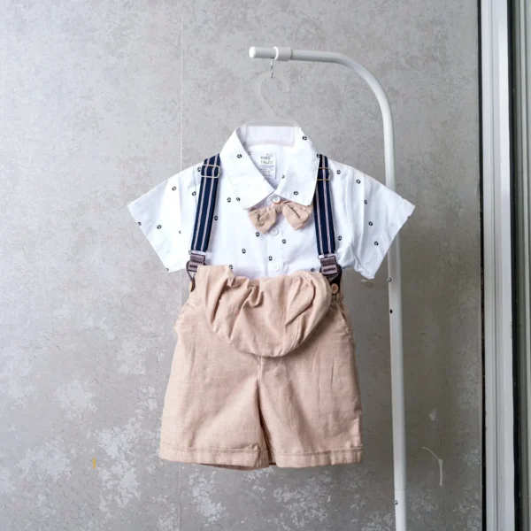 Baby boy’s cream-colored summer outfit with white short-sleeve shirt, tan suspender shorts, bow tie, and coordinating cap.