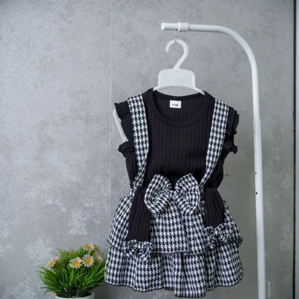 Baby girl’s 3-piece outfit with black ribbed top, houndstooth suspender skirt, large bow, and matching cap on hanger.