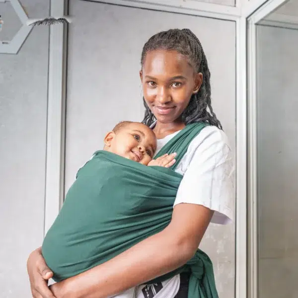 A young woman holds a baby close to her chest using a dark green baby wrap carrier, standing indoors against a light-colored wall with metal frames.
