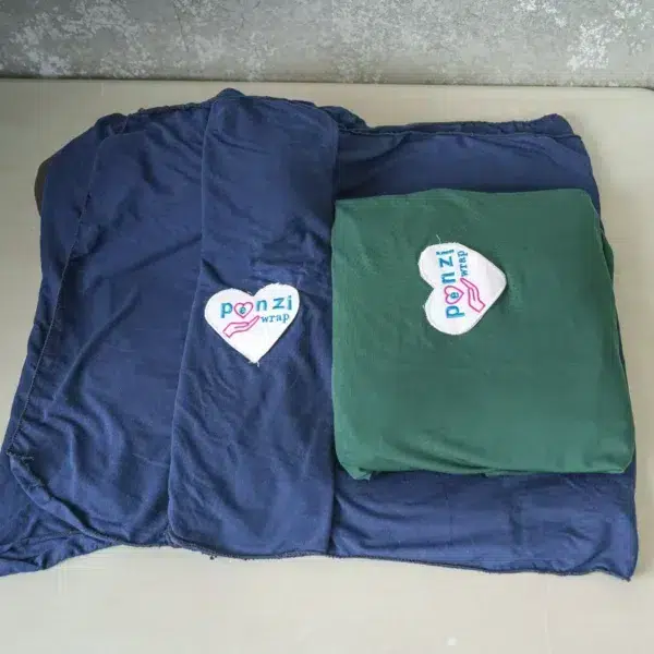 Three folded baby wrap carriers—light gray, navy blue, and dark green—are neatly displayed on a table, each with a heart-shaped logo reading "Ponzi Wrap".