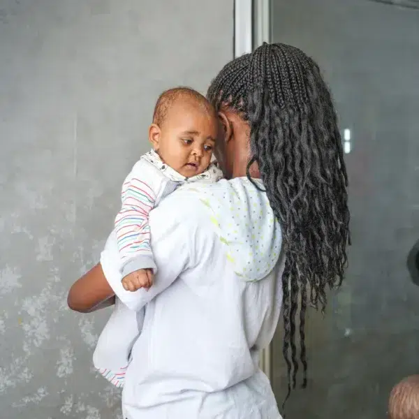 A smiling woman looks down at a baby held snugly in a light gray baby wrap carrier, both standing indoors with a soft, neutral background