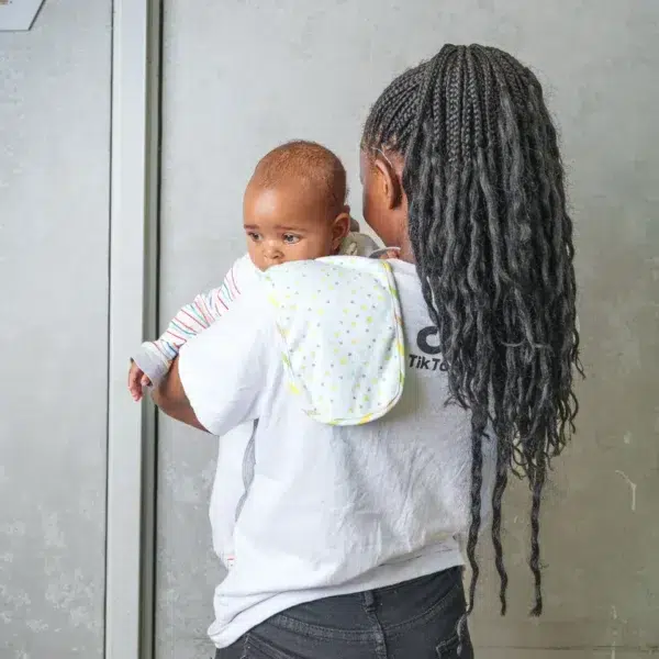 A smiling woman looks down at a baby held snugly in a light gray baby wrap carrier, both standing indoors with a soft, neutral background