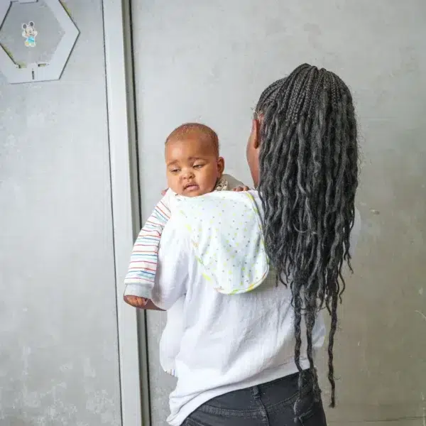 A smiling woman looks down at a baby held snugly in a light gray baby wrap carrier, both standing indoors with a soft, neutral background