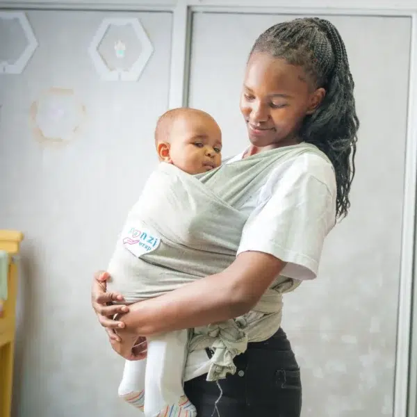 A smiling woman looks down at a baby held snugly in a light gray baby wrap carrier, both standing indoors with a soft, neutral background