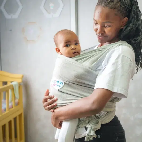 A smiling woman looks down at a baby held snugly in a light gray baby wrap carrier, both standing indoors with a soft, neutral background