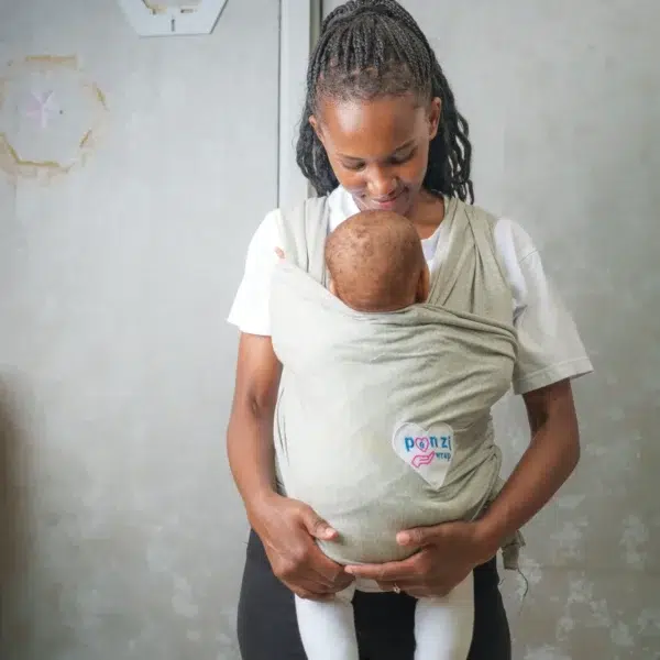 A smiling woman looks down at a baby held snugly in a light gray baby wrap carrier, both standing indoors with a soft, neutral background