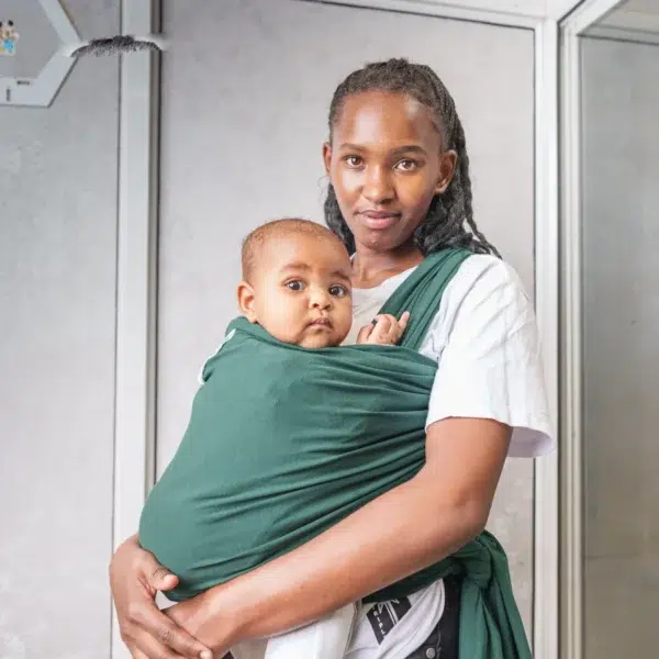 A young woman holds a baby close to her chest using a dark green baby wrap carrier, standing indoors against a light-colored wall with metal frames.