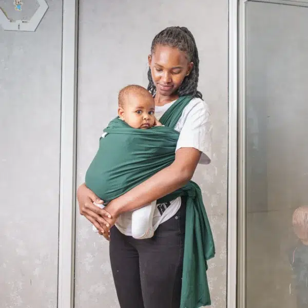A young woman holds a baby close to her chest using a dark green baby wrap carrier, standing indoors against a light-colored wall with metal frames.