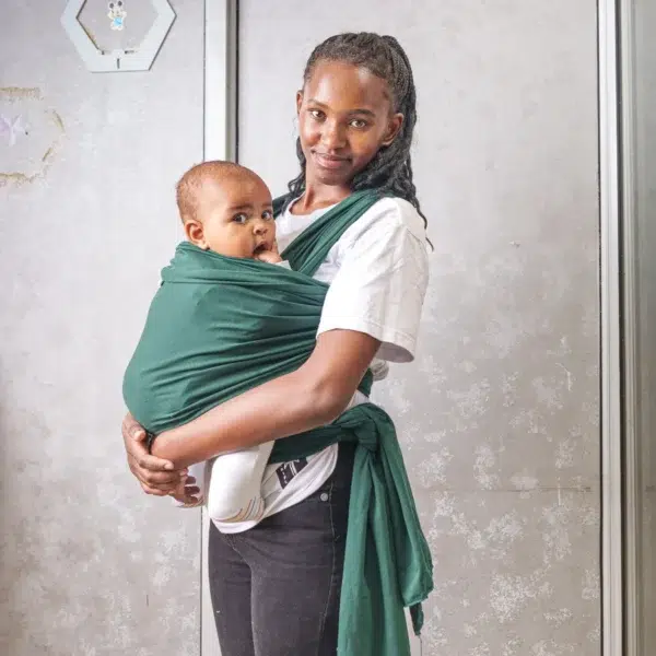 A young woman holds a baby close to her chest using a dark green baby wrap carrier, standing indoors against a light-colored wall with metal frames.