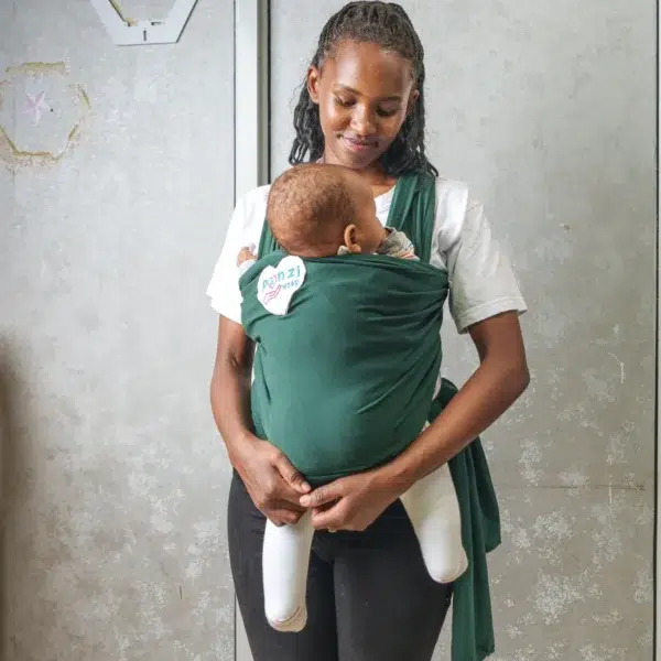 A young woman holds a baby close to her chest using a dark green baby wrap carrier, standing indoors against a light-colored wall with metal frames.
