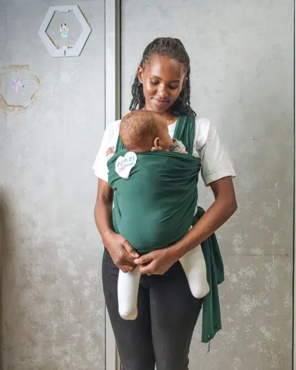 A young woman holds a baby close to her chest using a dark green baby wrap carrier, standing indoors against a light-colored wall with metal frames.
