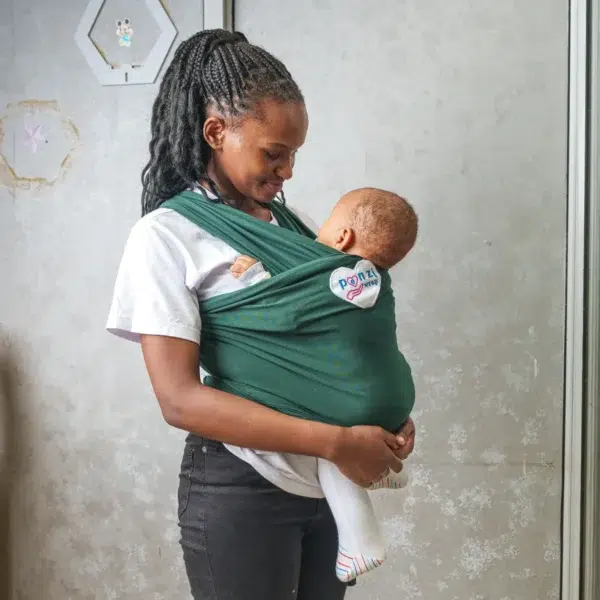 A young woman holds a baby close to her chest using a dark green baby wrap carrier, standing indoors against a light-colored wall with metal frames.
