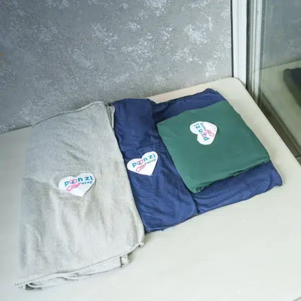 Three folded baby wrap carriers—light gray, navy blue, and dark green—are neatly displayed on a table, each with a heart-shaped logo reading "Ponzi Wrap".