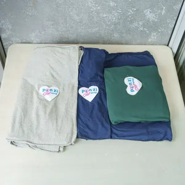 Three folded baby wrap carriers—light gray, navy blue, and dark green—are neatly displayed on a table, each with a heart-shaped logo reading "Ponzi Wrap".