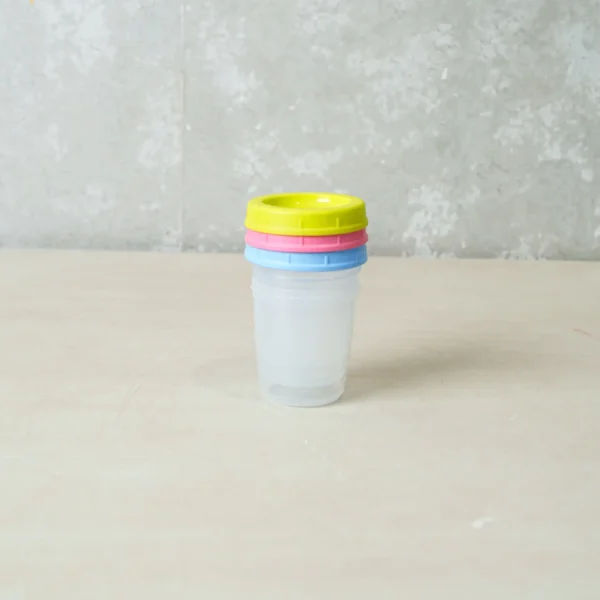 Three transparent baby food storage containers stacked without lids, alongside three colorful lids (yellow, pink, and blue) placed separately on a flat surface.