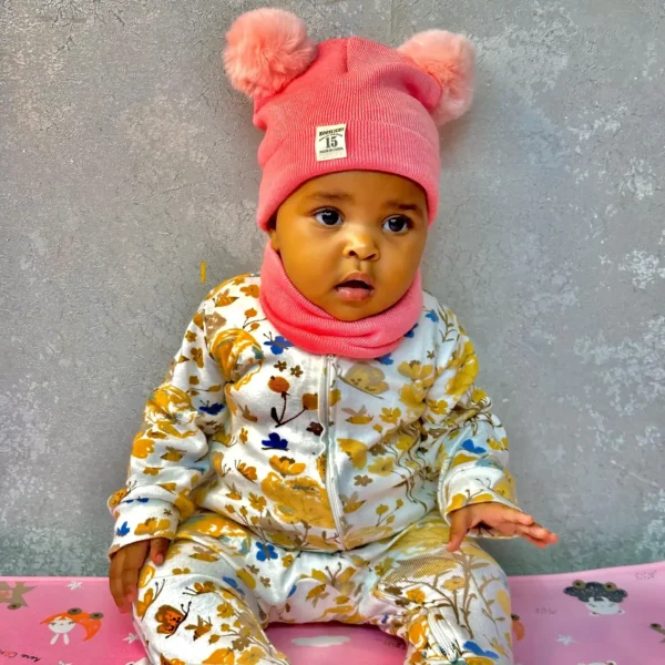 A baby wearing a pink beanie hat with two fluffy pom-poms and a matching neck warmer, dressed in a floral jumpsuit, sitting on a pink mat with cartoon prints.