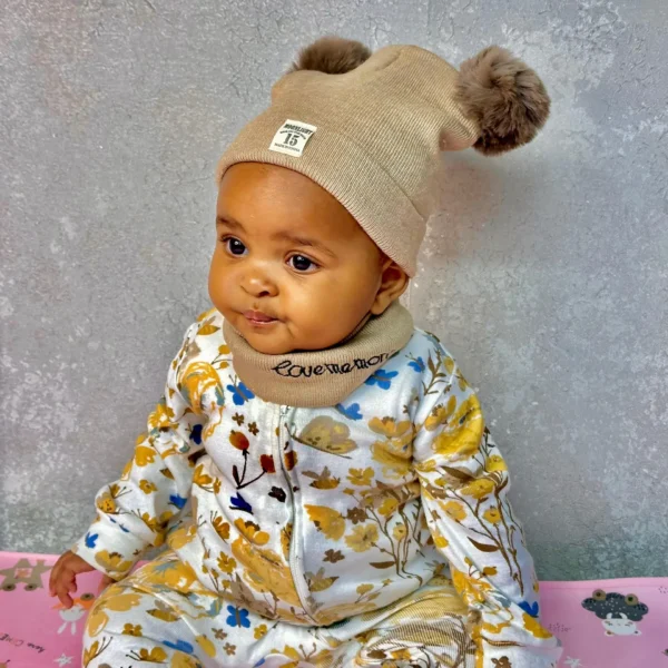 A baby wearing a beige beanie hat with two fluffy pom-poms and a matching neck warmer, sitting against a textured grey background, dressed in a floral jumpsuit.