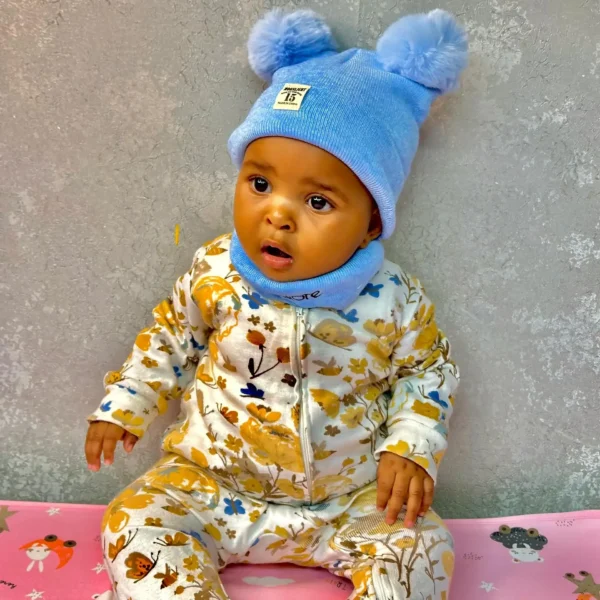 A baby wearing a light blue beanie hat with fluffy pom-poms and a matching neck warmer, sitting on a pink mat with playful designs, dressed in a floral jumpsuit.