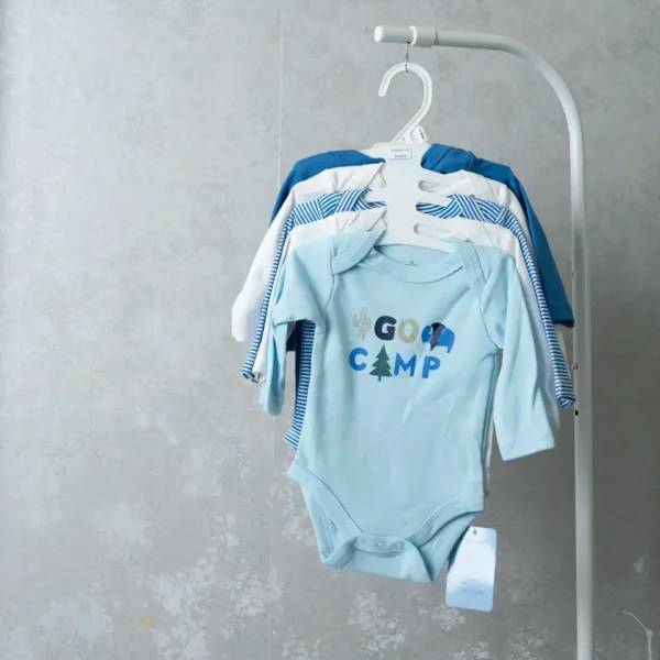 A set of blue-themed baby bodysuits on a white hanger, with the front onesie in light blue displaying camping-themed graphics and the text “Go Camp.