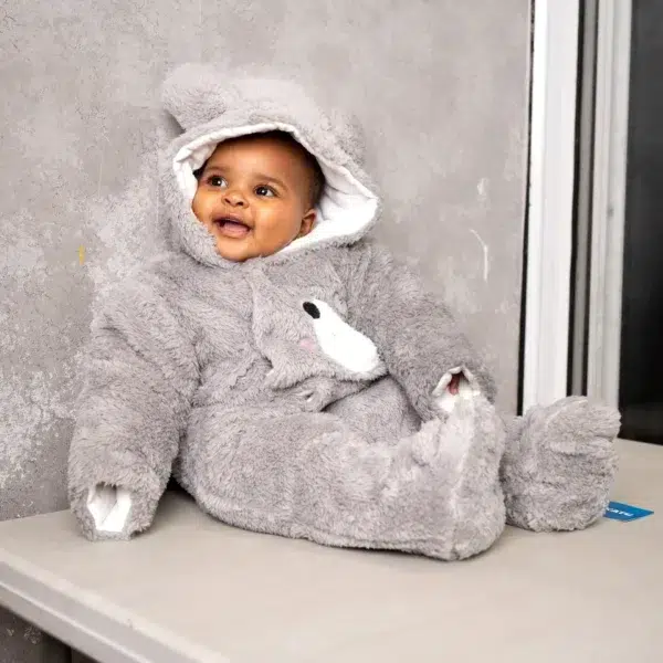Child sitting on a blanket in a hooded fleece romper, holding a soft toy, with natural light illuminating the scene.