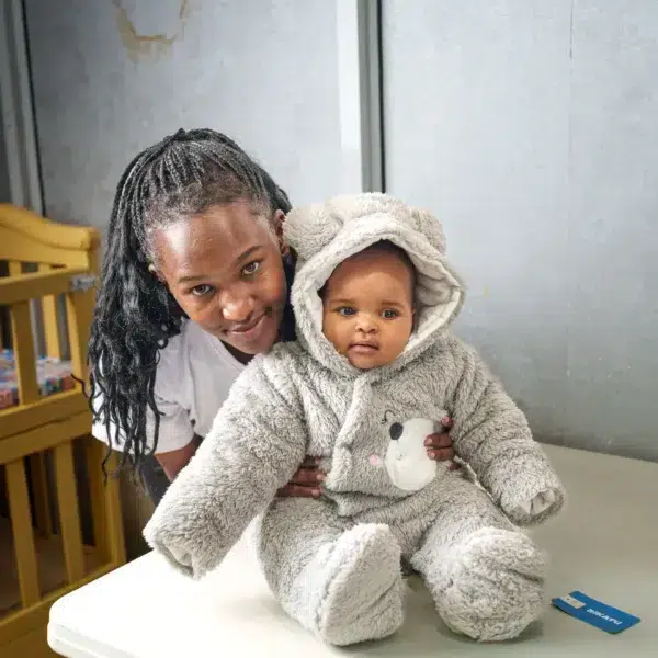Child sitting on a blanket in a hooded fleece romper, holding a soft toy, with natural light illuminating the scene.
