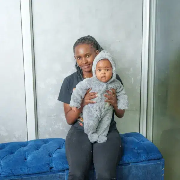 A woman sitting on a blue tufted bench holding a baby dressed in a gray fleece romper with a hood, in front of a light gray wall.