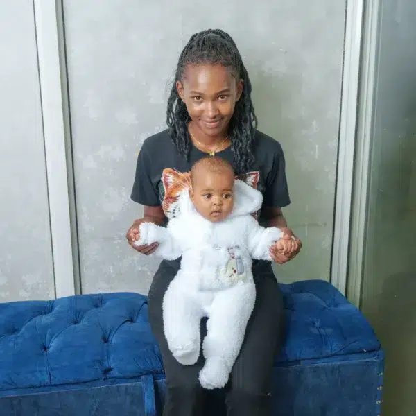 A woman sitting on a blue tufted bench holding a baby wearing a white fleece romper with a hood and elephant design, in front of a gray wall.