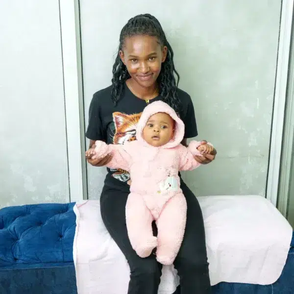 A woman sitting on a blue bench covered partly with a pink cloth, holding a baby dressed in a pink fleece romper with a hood and animal design, against a gray wall background.
