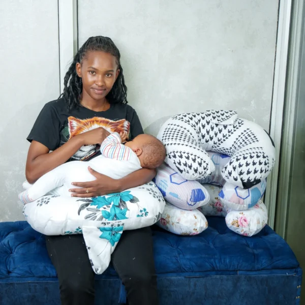 A mother breastfeeding her baby using a white and blue floral nursing pillow while another U-shaped nursing pillow with black-and-white patterns is placed beside her on a blue velvet seat.