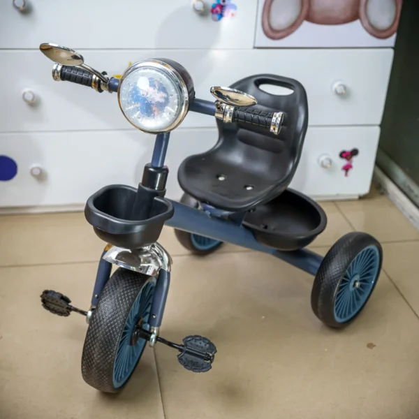hildren’s tricycle with a black seat, blue wheels, and handlebar mirrors, displayed indoors on a tiled floor near a white dresser with colorful decor.